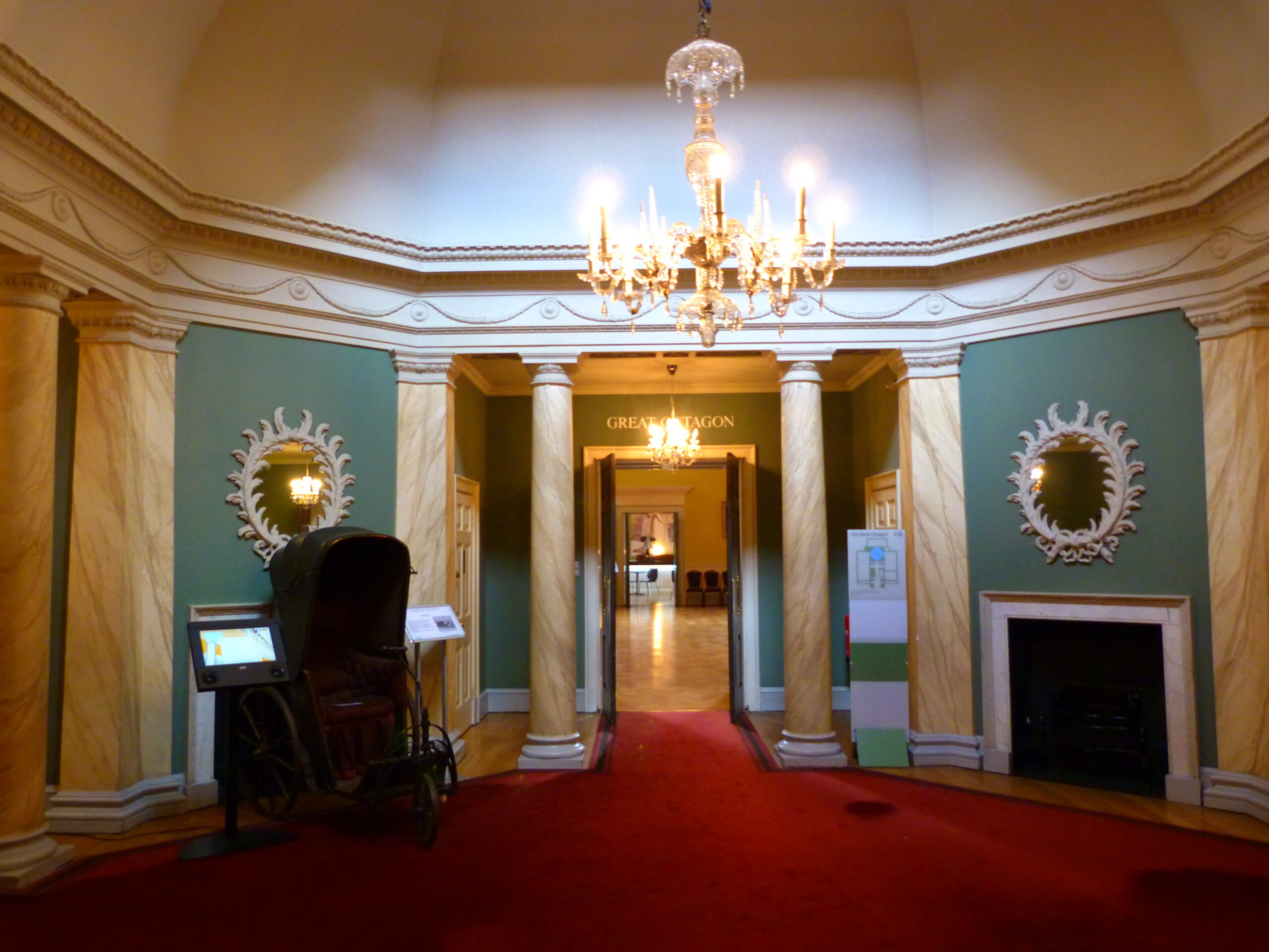 The entrance hall of Bath's Assembly Rooms, with marble-effect columns, a crystal chandelier, and an original sedan chair on display