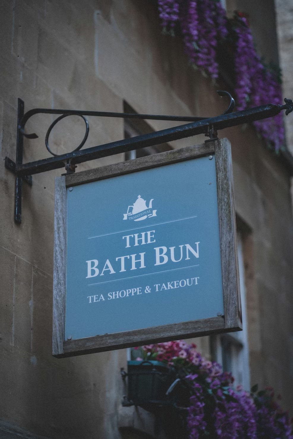 The Bath Bun Tea Shoppe sign on a Georgian stone wall, with purple wisteria trailing above